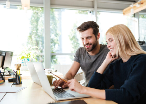 Image of happy young colleagues sitting in office coworking using laptop computers. Looking aside.
